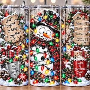 Peut inclure: Un gobelet cylindrique orné d'un bonhomme de neige emmêlé dans des guirlandes lumineuses colorées, une écharpe à carreaux rouges et un haut-de-forme. Le motif inclut des panneaux en bois avec le texte Just trying to keep it together, Coffee, Patience, Sanity, Christmas et Hot Mess Express.