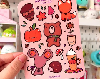 以下が含まれることがあります： 秋をテーマにしたイラストが描かれたピンク色のステッカーシート。シートには、キツネ、クマ、リンゴ、キノコ、その他の秋をテーマにした画像が描かれています。「Autumn Forest Sticker Sheet」の文字が上部にあります。アーティスト名「AlwaysOddity」が下部にあります。
