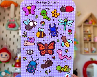 以下が含まれることがあります： 紫色のギンガムチェックの背景に、カラフルな漫画の昆虫と「Garden Critters」と「Always Oddity」の文字が描かれたステッカーシート。 蝶、蜂、てんとう虫などが含まれています。