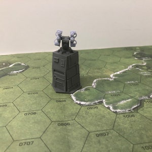 May include: A grey 3D printed model of a futuristic tower with four small, light grey, cylindrical turrets on top. The tower is sitting on a green hexagonal game board with white numbers.