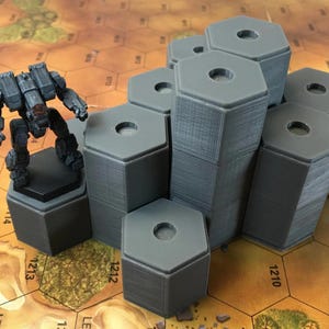 Könnte beinhalten: Ein Satz grauer sechseckiger Plastiksockel für Tabletop-Spiele. Die Sockel sind pyramidenförmig gestapelt, wobei der größte Sockel unten und der kleinste oben liegt. Die Sockel haben jeweils ein Loch in der Mitte.