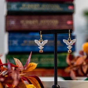 May include: A pair of gold-toned hoop earrings featuring white owl charms with spread wings. Each owl holds a small tan envelope with a red heart detail in its talons. The earrings are displayed on a black T-shaped stand against a blurred background.
