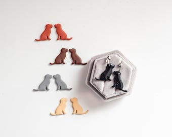 May include: A set of dog-shaped earrings in various colors, including black, brown, gray, and beige. The earrings are displayed on a white surface with a small gray box containing a pair of black dog earrings.
