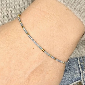 May include: A delicate bracelet made with small grey and gold beads. The bracelet is worn on a person's wrist.