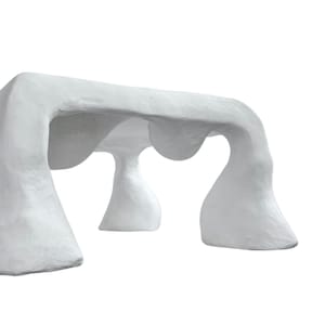 Könnte beinhalten: Ein weißer, skulpturaler Tisch mit organischem, höhlenartigem Design. Der Tisch hat eine rechteckige Oberseite, die von mehreren abgerundeten, säulenartigen Beinen getragen wird. Die Gesamtästhetik ist glatt und modern, mit Fokus auf Form und Textur.
