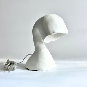 May include: A white, sculptural table lamp with a curved, organic form. The lamp has a rounded base that tapers into a slender neck, then curves upward to a flared shade. A grey power cord with a plug extends from the base.