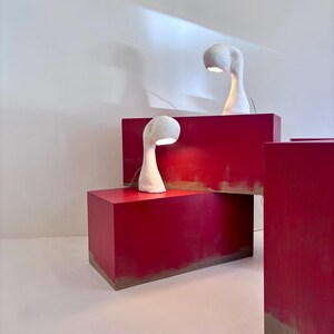 May include: Two white, sculptural table lamps with curved necks and rounded shades are displayed on bright red rectangular blocks. The lamps are lit, casting a warm glow. The blocks are arranged in a studio setting with a white background.