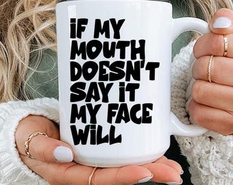May include: White ceramic mug with the text "IF MY MOUTH DOESN'T SAY IT MY FACE WILL" in black, bold letters. The mug has a large handle and is held by a person wearing a green shirt and a white sweater.