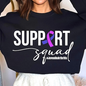 Puede incluir: Sudadera negra con texto blanco que dice "SUPPORT squad #JuvenileArthritis" y una cinta morada y azul.