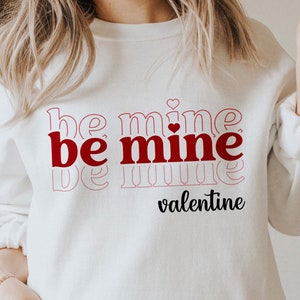 May include: White sweatshirt with the text "be mine be mine be mine" in red and pink with a small red heart. The text "valentine" is written in black below the main text.