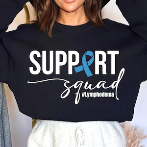 Puede incluir: Sudadera negra con el texto "SUPPORT squad #Lymphedema" y una cinta azul.