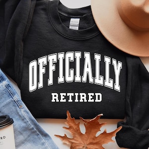 May include: Black sweatshirt with white text that reads "Officially Retired". The sweatshirt is laid out on a white surface with a pair of blue jeans, a coffee cup, and a brown hat.