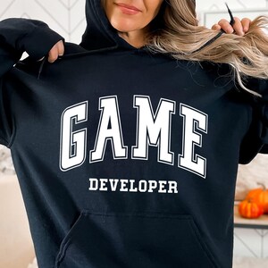 Pode incluir: Camisola de moletom com capuz preta com texto branco que diz "GAME DEVELOPER" em uma fonte universitária.