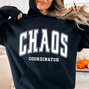 May include: Black hoodie with the words "CHAOS COORDINATOR" in white block letters. The hoodie has a front pocket and a drawstring hood. The design is centered on the chest.
