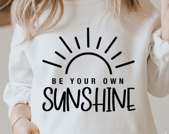 Può includere: Felpa bianca con un grafico di sole nero e il testo "Sii la tua propria luce del sole".