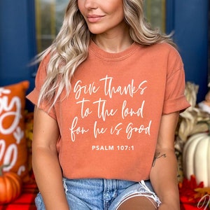 May include: A woman wearing a burnt orange t-shirt with the text "Give thanks to the Lord for he is good, Psalm 107:1" in white. She is wearing a beige beanie and blue denim shorts.