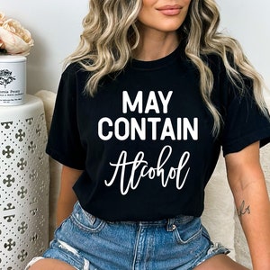 May include: Black t-shirt with the text "MAY CONTAIN Alcohol" in white, uppercase letters above and cursive below. The shirt is worn with denim shorts. A white side table with a floral arrangement and a candle is in the background.