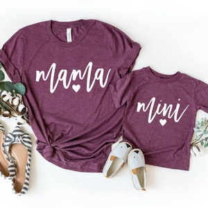 Puede incluir: Dos camisetas color granate con texto blanco. La camiseta más grande dice "mama" con dos corazones pequeños. La camiseta más pequeña dice "mini" con un corazón pequeño.