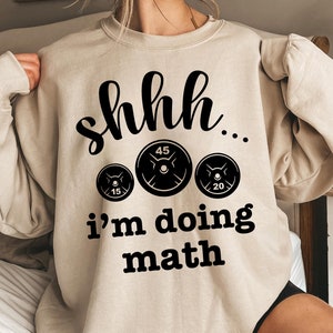 Könnte beinhalten: Ein beiger Pullover mit schwarzem Text, der "shhh... i'm doing math" lautet und drei schwarze Gewichtsplatten mit den Zahlen 15, 45 und 20 zeigt.