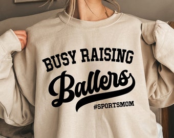 Op de afbeelding: Beige sweatshirt met de zwarte tekst "BUSY RAISING Ballers" en "#SPORTSMOM" eronder. De sweatshirt heeft een ronde hals.