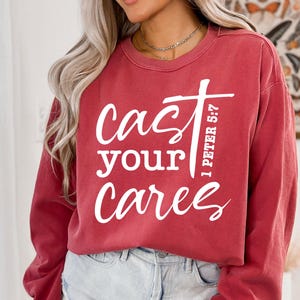 Puede incluir: Sudadera roja de manga larga con el texto "cast your cares" y el versículo bíblico "1 Pedro 5:7" en blanco.