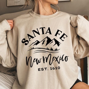 May include: Beige sweatshirt with a black graphic design featuring the words "SANTA FE" above a mountain range and the words "New Mexico" below. The text "EST. 1610" is printed below the state name. The sweatshirt is a neutral color.