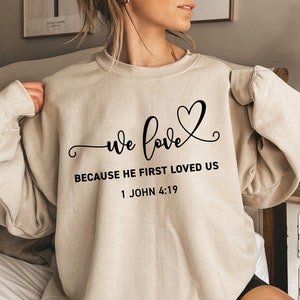 May include: A beige sweatshirt with the text "we love because he first loved us 1 John 4:19" in black lettering. The text is accompanied by a stylized heart.