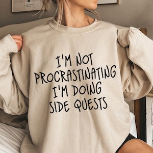 Könnte beinhalten: Ein beiger Pullover mit dem Aufdruck "I'm not procrastinating I'm doing side quests" in Schwarz.