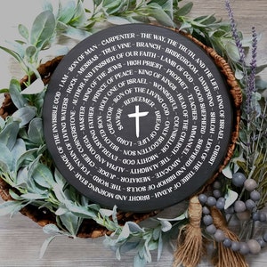 May include: A black circular wooden sign with white text in a spiral pattern. The text includes names and titles of Jesus Christ. The sign is surrounded by greenery and a strand of gray beads.