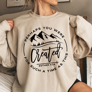 May include: A beige sweatshirt with a black graphic design that says "Perhaps you were created for such a time as this" with a mountain silhouette and the scripture reference "Esther 4:14".