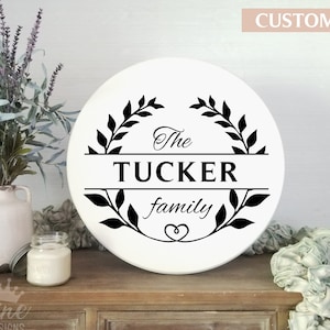 May include: White round wooden sign with black lettering that reads "The Tucker family" surrounded by a wreath of leaves. The sign is on a wooden shelf with a vase of lavender and a candle.