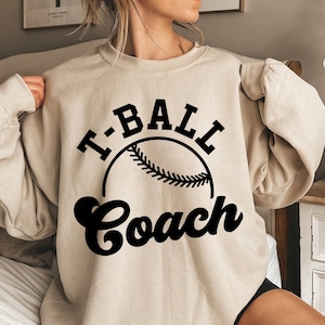 Könnte beinhalten: Ein beiger Pullover mit dem Text "T-BALL COACH" in schwarzen Buchstaben. Eine schwarze Baseballgrafik befindet sich mittig über dem Text.