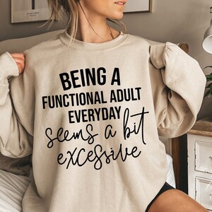 May include: Beige crewneck sweatshirt with the text "BEING A FUNCTIONAL ADULT EVERYDAY seems a bit excessive" in black. The sweatshirt is a neutral color and has long sleeves. The text is in a bold, sans-serif font.