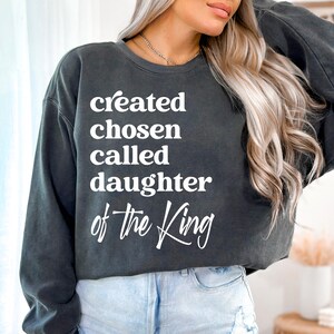 May include: A dark grey sweatshirt with the text "created chosen called daughter of the King" in white.