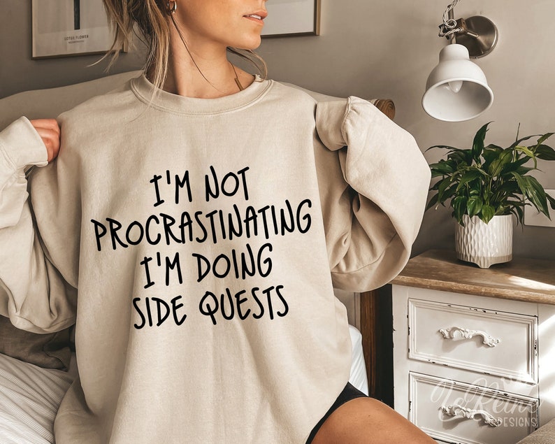 May include: A beige sweatshirt with the text "I'm not procrastinating I'm doing side quests" printed in black.