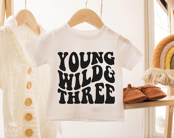 May include: White t-shirt with black text that reads "Young Wild & Three" in a retro font style.