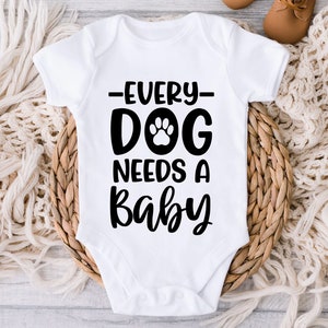 Op de afbeelding: Witte baby romper met zwarte tekst die "Every Dog Needs A Baby" luidt met een poot afdruk grafiek.