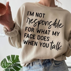 Könnte beinhalten: Beiges Sweatshirt mit schwarzem Text: "I'M NOT responsible FOR WHAT MY FACE DOES WHEN YOU talk". Das Sweatshirt hat lange Ärmel und einen Rundhalsausschnitt. Das Le Reine Designs-Logo befindet sich unten.
