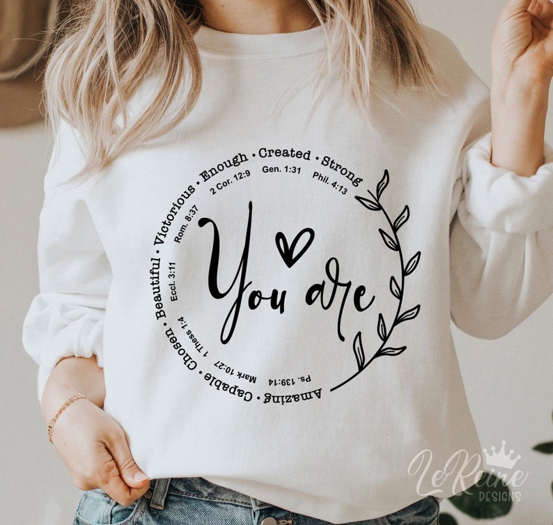 May include: White sweatshirt with a black circle design that says "You are" with a heart in the middle. The circle is surrounded by a wreath of leaves and the words "Amazing Capable Chosen Beautiful Victorious Enough Created Strong" with bible verses listed below each word.
