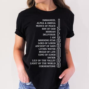 Op de afbeelding: Zwart T-shirt met witte tekst die "JESUS" verticaal langs de rechterkant van het shirt loopt. De tekst bestaat uit verschillende titels en beschrijvingen van Jezus, waaronder "Emmanuel", "Alfa & Omega", "Prins van de Vrede", "Zoon van God", "Messias", "Verlosser", "Ik ben", "Morgenster", "Heer der Heren", "Oude van Dagen", "Levend Water", "Brot des Lebens", "Koning der Koningen", "Redder", "Lelie van de Vallei", "Licht van de Wereld" en "Hoeksteen".