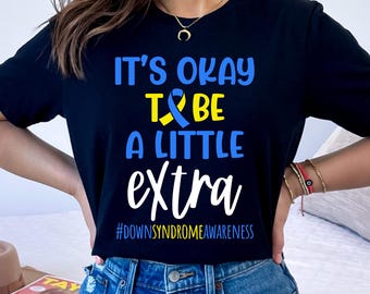 May include: Black t-shirt with white text that reads "It's okay to be a little extra #downsyndromeawareness". A blue and yellow ribbon is incorporated into the text. The person wearing the shirt is standing in front of a brown surface.