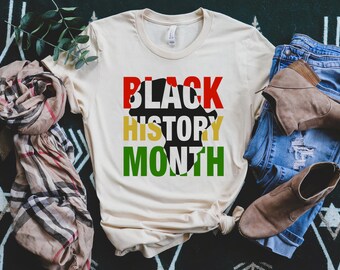 May include: A beige t-shirt with the text "Black History Month" in black, red, yellow, and green letters. The text is stylized to look like an outline of the African continent.