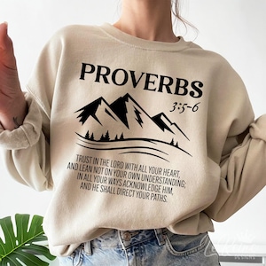 May include: Beige sweatshirt featuring a black mountain graphic and the text "Proverbs 3:5-6" and a quote from the Bible: "Trust in the Lord with all your heart, and lean not on your own understanding; In all your ways acknowledge him, and he shall direct your paths."
