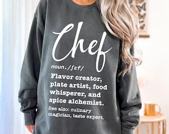 May include: A dark gray sweatshirt with a white graphic that reads "Chef" and defines the word as "Flavor creator, plate artist, food whisperer, and spice alchemist." The text also includes "See also: culinary magician, taste expert."