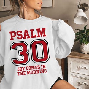 Pode incluir: Camisola branca com letras vermelhas que dizem "PSALM 30 JOY COMES IN THE MORNING".