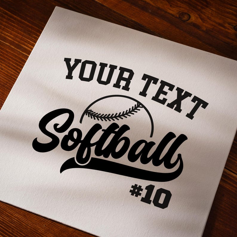 Pu&ograve; includere: Una tela bianca con un design di softball nero. Il testo recita "YOUR TEXT" sopra una grafica di softball, con "Softball" in un carattere script e il numero "10" sotto.