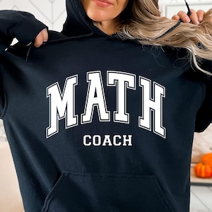 May include: Black hooded sweatshirt with the words "MATH COACH" in white block letters. The sweatshirt has a front pocket and a drawstring hood. The text is in a collegiate style font.