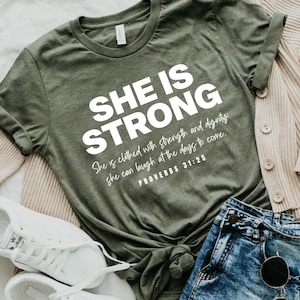 May include: A green t-shirt with the text "SHE IS STRONG" in white. Below the text is a quote from Proverbs 31:25, "She is clothed with strength and dignity: she can laugh at the days to come."