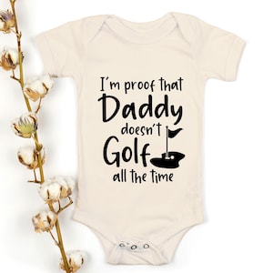 Puede incluir: Un body blanco para bebé con texto negro que dice "Soy la prueba de que papá no juega al golf todo el tiempo". El texto está en una fuente divertida e incluye un pequeño gráfico de un hoyo de golf y una bandera.
