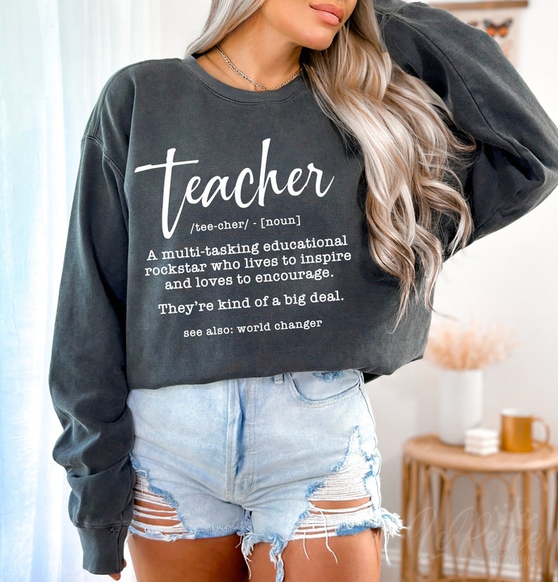 May include: A gray sweatshirt with the word "teacher" written in white. The text below defines the word as a "multi-tasking educational rockstar who lives to inspire and loves to encourage." The text also says "They're kind of a big deal." and "see also: world changer."